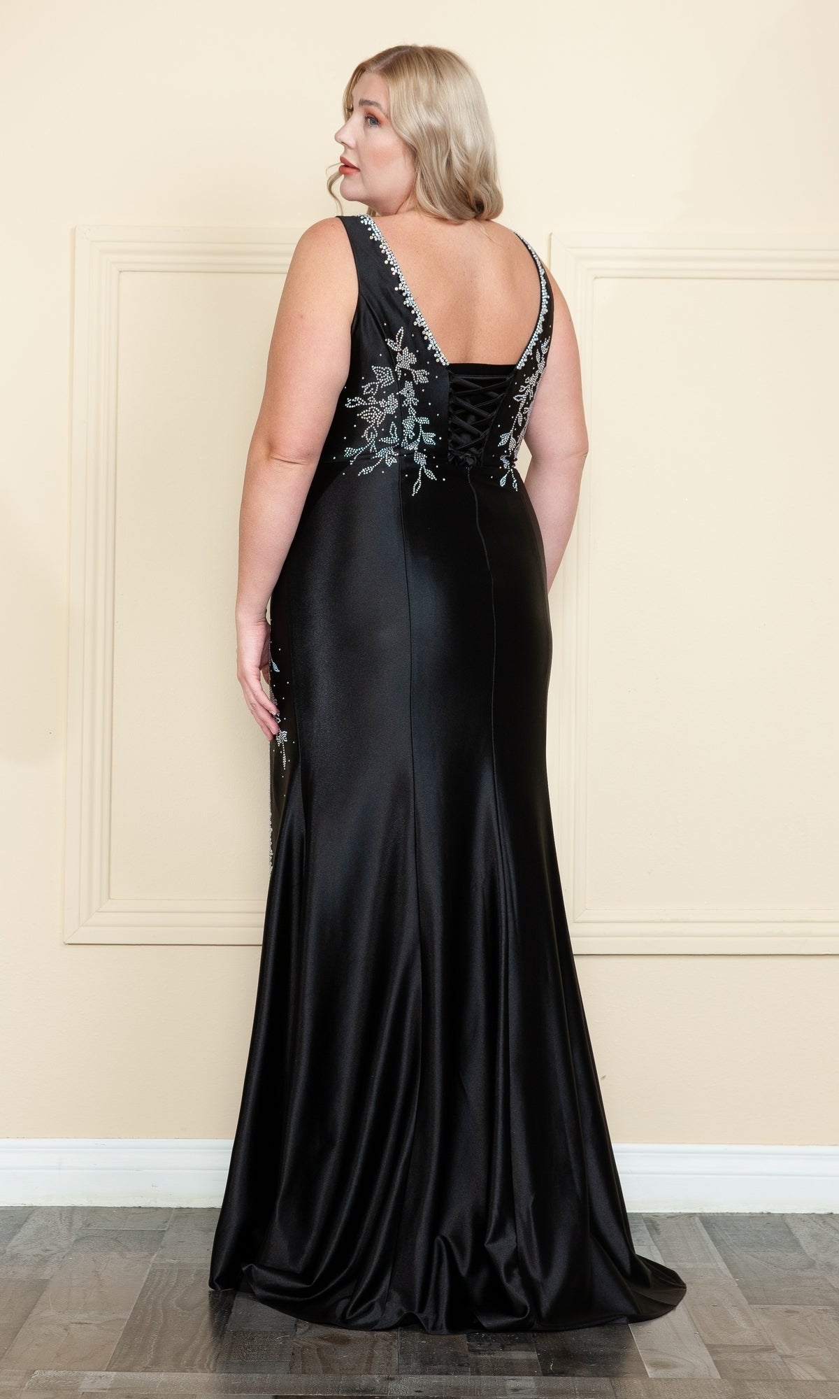 A woman with blonde hair faces away, wearing the Plus-Size Long Spandex Prom Dress with Beads W1132, featuring silver details on the back, standing indoors against a light-colored wall.